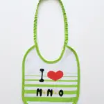 Bavetta neonato bianca con bordi verde lime, laccetto elastico arricciato e scritta I Love Nonno con cuore rosso su righe verdi e bianche.