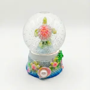 Palla di neve con tartaruga marina carapace rosa corallo e pinne verde lime, base decorata con conchiglie, alghe e perla bianca