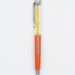 Penna a sfera arancione metallizzato con sezione trasparente e cristalli gialli, gommino arancione, clip cromata, scritta BAHJA incisa sul corpo