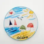 Piatto ceramica con mare azzurro, barche a vela blu e rossa, ombrelloni verde e rosso e scritta Calabria sulla sabbia gialla