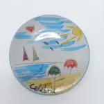 Piatto ceramica rotondo con scena balneare: mare azzurro, barche a vela rossa e verde, ombrelloni, sole e scritta Calabria in nero
