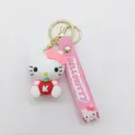Portachiavi Hello Kitty: ciondolo bianco con fiocco rosa e mela rossa lettera K, cinturino in gomma rosa con scritta HELLO KITTY e moschettone dorato.