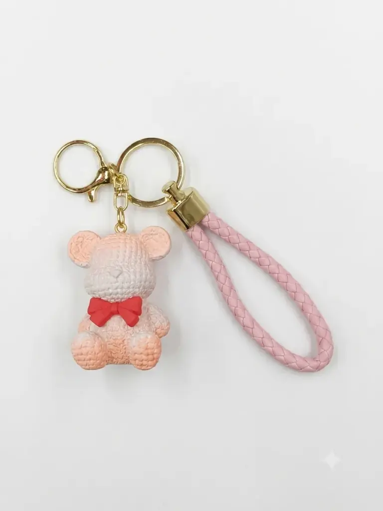 Portachiavi orsetto tridimensionale rosa-pesca e bianco con texture a maglia, fiocco rosso al collo, cordino intrecciato rosa e anelli dorati