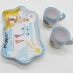 Vassoio sagomato bordo azzurro ondulato con scritta Calabria nera e due tazzine caffè ceramica smaltata, motivi rossi gialli e verdi su fondo bianco