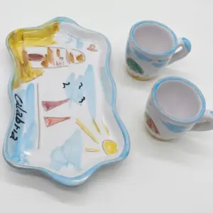 Vassoio sagomato bordo azzurro ondulato con scritta Calabria nera e due tazzine caffè ceramica smaltata, motivi rossi gialli e verdi su fondo bianco
