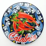 Sottopentola rotondo ceramica, tre peperoncini rossi su fondo blu scuro con volute bianche e scritta Calabria in corsivo rosso